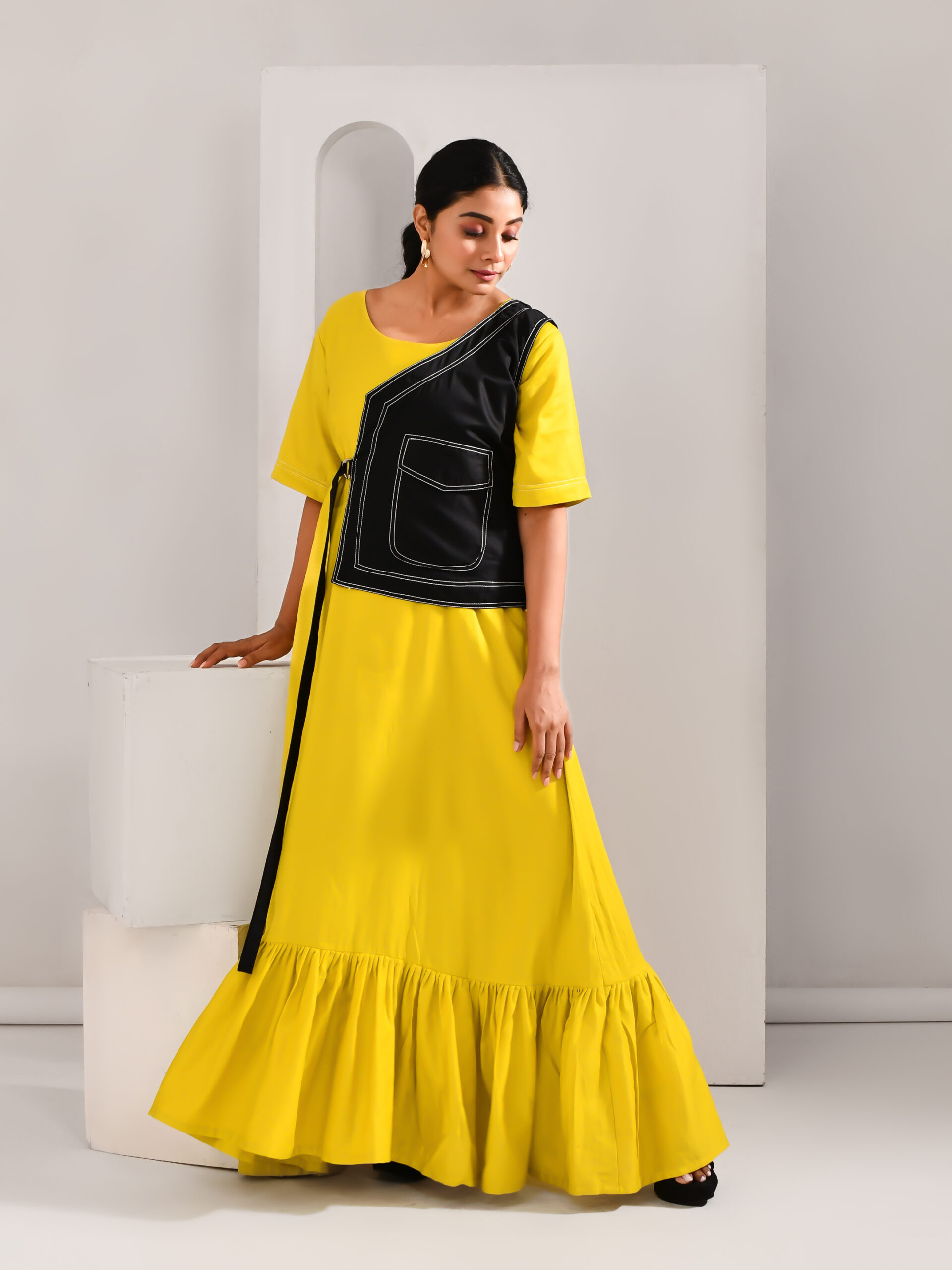 “Golden Muslin golden yellow blended cotton maxi dress paired with black vest-style jacket featuring waist tie-up belt.”
